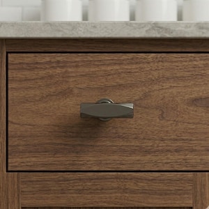 May include: Close-up of a wooden drawer with a dark metal handle. The drawer is made of brown wood with visible grain. The handle is a rectangular shape with rounded edges and a matte finish. The drawer is part of a larger cabinet.