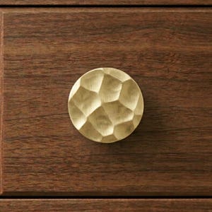 May include: A round, gold-colored cabinet knob with a textured, hammered surface. The knob is mounted on a dark brown wooden drawer front, showcasing a contrast in textures and colors. This hardware adds a touch of elegance to furniture.