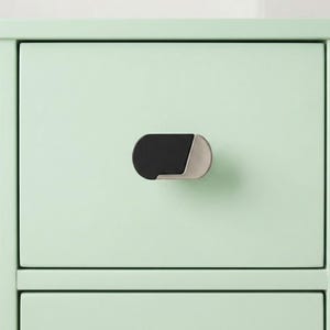 May include: Close-up of a mint green cabinet drawer with a black and silver oval-shaped handle. The handle is mounted horizontally on the drawer front, providing a modern and minimalist aesthetic.
