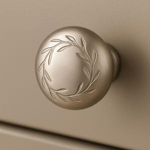 May include: A brushed nickel cabinet knob with a circular leaf design. The knob is round with a small stem and is mounted on a neutral-toned surface. The leaf design is etched into the surface of the knob.