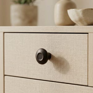 May include: Close-up of a black cabinet knob on a light beige drawer. The knob is round with a flat face and a short stem. The drawer has a textured, linen-like appearance. The background includes a ceramic vase and a small bowl.