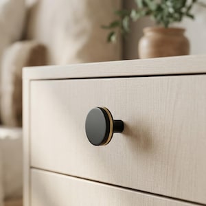May include: A black cabinet knob with a gold accent ring is attached to a light-colored wooden drawer. The knob is round and has a simple, modern design. The drawer is part of a larger piece of furniture, possibly a dresser or nightstand.