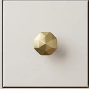 May include: A gold-colored, geometric cabinet knob with an octagonal shape. The knob is mounted on a white painted wooden surface. The knob has a faceted design, reflecting light. The overall aesthetic is modern and minimalist, suitable for furniture hardware.