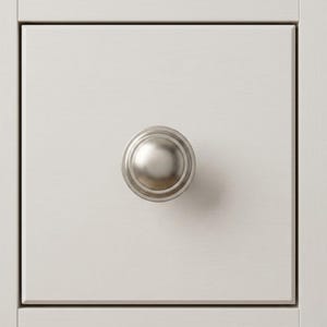 May include: Close-up of a white painted drawer with a brushed nickel knob. The knob is round with a slightly domed center and a circular base. The drawer has a simple, clean design.