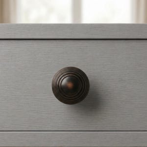 May include: Close-up of a gray wooden drawer with a dark bronze, round knob. The knob has a series of concentric rings, creating a textured appearance. The drawer's surface has a visible wood grain.