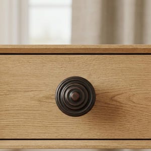 May include: Close-up of a wooden drawer with a dark bronze, circular knob. The knob has a layered design, and the drawer is made of light-colored wood with a visible grain. The background is blurred, suggesting a room setting.