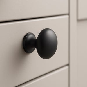 May include: Close-up of a black, oval-shaped cabinet knob with a circular base. The knob is mounted on a light beige cabinet drawer. The knob has a matte finish and is centered in the frame.