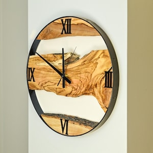 Könnte beinhalten: Eine runde Wanduhr mit schwarzem Metallrahmen und römischen Ziffern. Das Zifferblatt hat ein natürliches Holzmaserungsdesign und schwarze Zeiger. Die Uhr ist an einer weißen Wand befestigt.