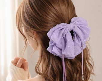 May include: A large lavender hair bow clip made of textured fabric with a subtle grid pattern and small raised dots. The bow features two long, thin lavender ribbons hanging down, each finished with a small, clear, teardrop-shaped crystal charm.
