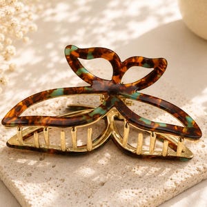 May include: A tortoiseshell-patterned hair claw clip with a butterfly design. The clip features shades of brown, gold, and teal. The hair accessory is resting on a textured, light-colored surface.