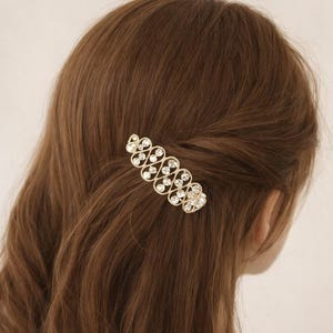 May include: A gold-toned hair clip with a decorative design of alternating clear, round rhinestones. The hair accessory is set against a backdrop of long, wavy brown hair. The clip is positioned to secure a section of hair.