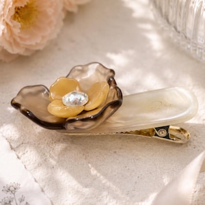 May include: A hair clip featuring a translucent brown and cream-colored flower ornament with a central clear rhinestone. The clip has a smooth, pearlescent off-white base and a gold-toned metal hinge mechanism, showcasing a refined and elegant design.