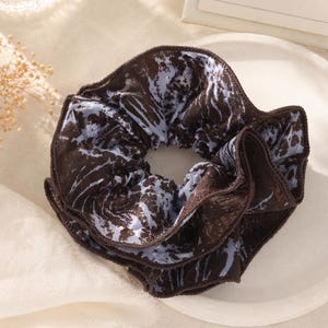 May include: A brown scrunchie with a marbled pattern in shades of blue and white. The hair accessory is made of a soft fabric and is designed to hold hair in place. The scrunchie is a stylish and practical hair accessory.
