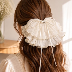 May include: A cream-colored hair accessory featuring layered, ruffled fabric in a fan shape. The piece is adorned with a central white pearl and a small clear crystal, with two thin ribbons hanging down from the center of the ruffled design.