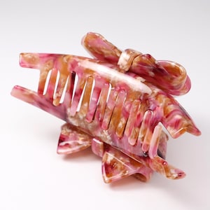 Pu&ograve; includere: Un fermaglio per capelli con motivo marmorizzato nei toni del rosa, bianco e ambra. L'accessorio presenta una serie di denti a incastro sui lati e un elemento decorativo a forma di fiocco fissato sulla parte superiore della struttura.
