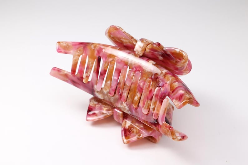 Pu&ograve; includere: Un fermaglio per capelli con motivo marmorizzato nei toni del rosa, bianco e ambra. L'accessorio presenta una serie di denti a incastro sui lati e un elemento decorativo a forma di fiocco fissato sulla parte superiore della struttura.