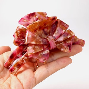 Pu&ograve; includere: Un fermaglio per capelli a forma di fiocco multistrato con finitura lucida. L'accessorio presenta un motivo marmorizzato nei toni del rosa scuro, bordeaux, bianco e ambra traslucido, dotato di denti a incastro per una presa sicura.