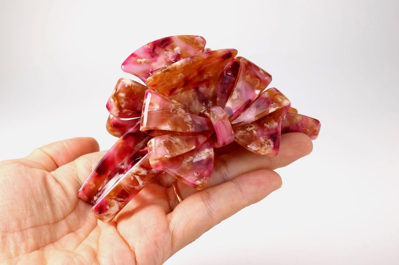 Pu&ograve; includere: Un fermaglio per capelli a forma di fiocco multistrato con finitura lucida. L'accessorio presenta un motivo marmorizzato nei toni del rosa scuro, bordeaux, bianco e ambra traslucido, dotato di denti a incastro per una presa sicura.