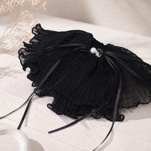 May include: A black ruffled collar accessory made of sheer, pleated fabric. The piece features a central decorative accent consisting of a single white pearl and a clear, faceted crystal, tied with thin black satin ribbons that drape downwards.