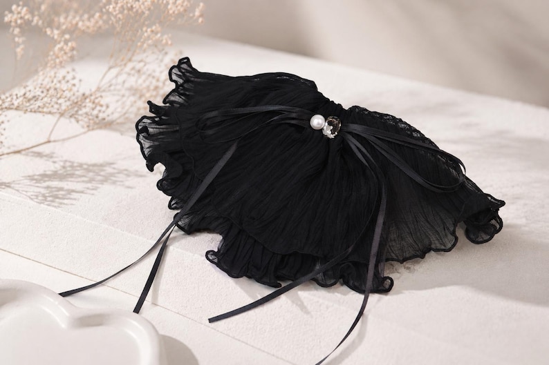 May include: A black ruffled collar accessory made of sheer, pleated fabric. The piece features a central decorative accent consisting of a single white pearl and a clear, faceted crystal, tied with thin black satin ribbons that drape downwards.