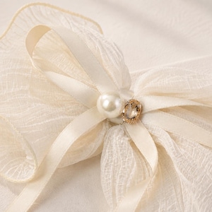 May include: A cream-colored hair bow crafted from sheer, textured fabric with ruffled edges. The center of the bow is adorned with a smooth white pearl and a small, oval-shaped faceted crystal set in a gold-toned metal frame.