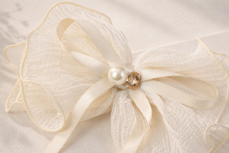 May include: A cream-colored hair bow crafted from sheer, textured fabric with ruffled edges. The center of the bow is adorned with a smooth white pearl and a small, oval-shaped faceted crystal set in a gold-toned metal frame.
