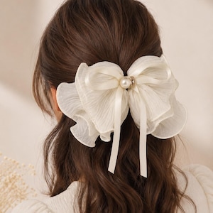 May include: A cream-colored hair bow made of layered, sheer, ruffled fabric. The center of the bow features a smooth, round white pearl bead accented by a small, clear, sparkling crystal stone, with two long, thin satin ribbons hanging down.