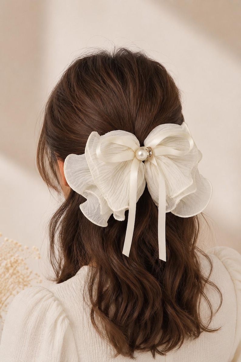 May include: A cream-colored hair bow made of layered, sheer, ruffled fabric. The center of the bow features a smooth, round white pearl bead accented by a small, clear, sparkling crystal stone, with two long, thin satin ribbons hanging down.