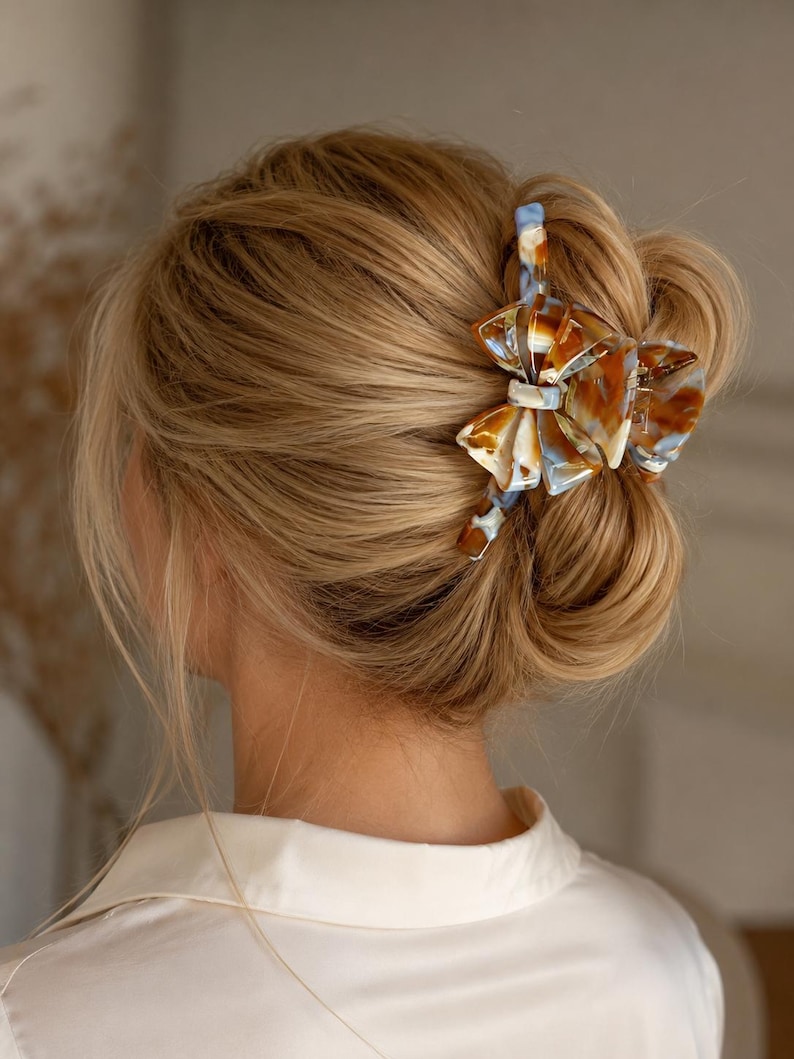 Pu&ograve; includere: Un fermaglio per capelli a forma di fiocco con un motivo marmorizzato nei toni del marrone, bianco e azzurro. Il fermaglio &egrave; realizzato in resina lucida e fissa i capelli biondi in uno chignon, mettendone in risalto i dettagli complessi.