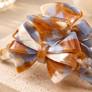 Pu&ograve; includere: Un fermaglio per capelli con un design a fiocco decorativo e un motivo marmorizzato nei toni del marrone, bianco e azzurro. L'accessorio ha una finitura lucida e un meccanismo a cerniera con denti a incastro per fissare i capelli.