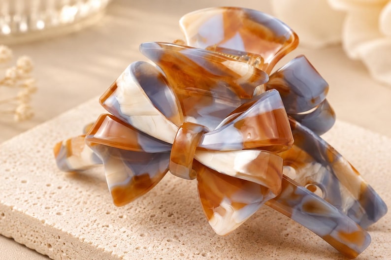 Pu&ograve; includere: Un fermaglio per capelli con un design a fiocco decorativo e un motivo marmorizzato nei toni del marrone, bianco e azzurro. L'accessorio ha una finitura lucida e un meccanismo a cerniera con denti a incastro per fissare i capelli.