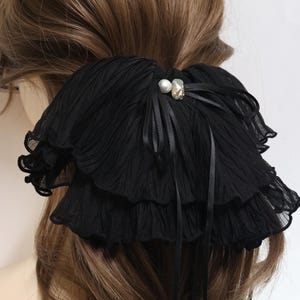 May include: A black hair accessory featuring layered, pleated fabric ruffles. The center is adorned with a small white pearl and a clear, faceted crystal bead, accented by thin black ribbon ties that drape downwards over the pleated material.