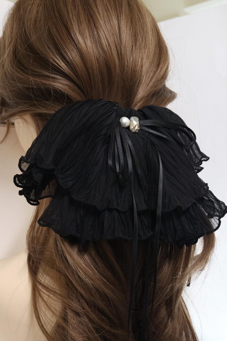 May include: A black hair accessory featuring layered, pleated fabric ruffles. The center is adorned with a small white pearl and a clear, faceted crystal bead, accented by thin black ribbon ties that drape downwards over the pleated material.