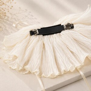 May include: A cream-colored hair accessory featuring a wide, pleated fabric ruffle attached to a black metal barrette clip. The fabric has a crinkled texture and is accented with two thin, matching cream-colored ribbons hanging from the sides.