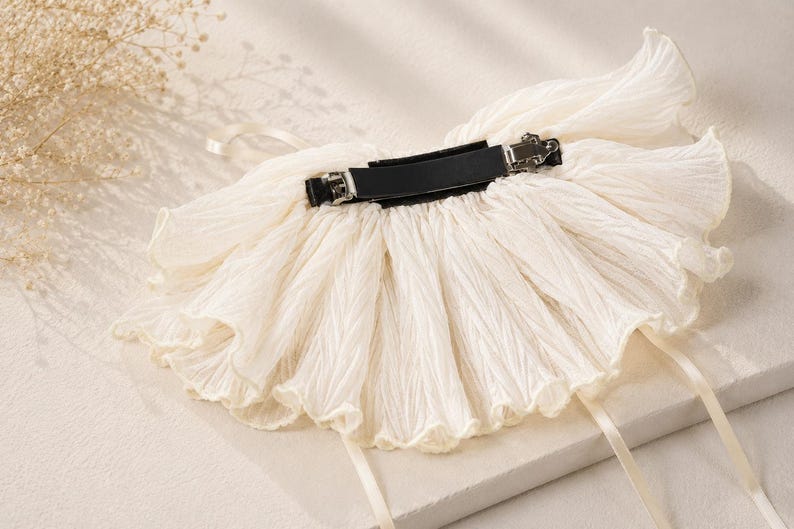 May include: A cream-colored hair accessory featuring a wide, pleated fabric ruffle attached to a black metal barrette clip. The fabric has a crinkled texture and is accented with two thin, matching cream-colored ribbons hanging from the sides.