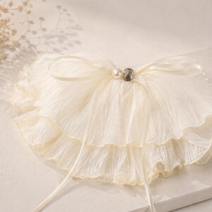 May include: A cream-colored, double-layered ruffled collar made of crinkled fabric. The collar features a thin ribbon tie at the center, accented with a single white pearl bead and a small, dark, faceted gemstone bead at the neckline.