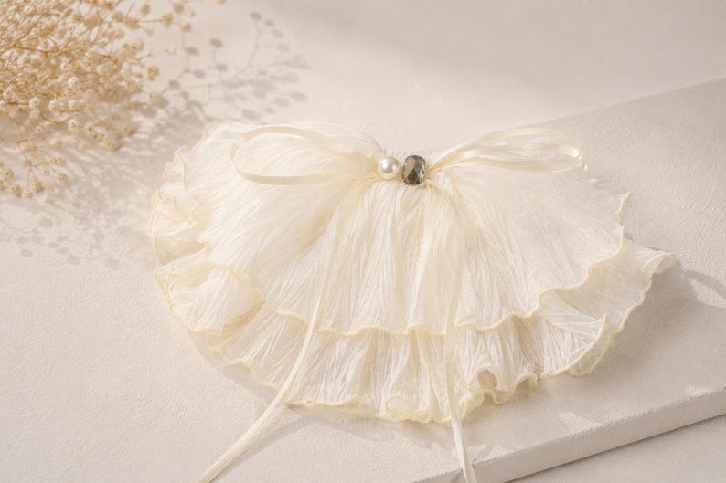 May include: A cream-colored, double-layered ruffled collar made of crinkled fabric. The collar features a thin ribbon tie at the center, accented with a single white pearl bead and a small, dark, faceted gemstone bead at the neckline.