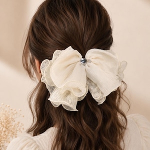May include: A large, off-white hair bow made of layered, sheer, and lace-trimmed fabric. The bow features a central, small, dark, metallic-toned circular accent piece that secures the gathered material, creating a voluminous, ruffled appearance.