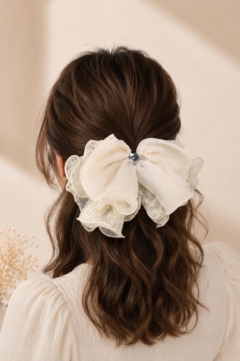 May include: A large, off-white hair bow made of layered, sheer, and lace-trimmed fabric. The bow features a central, small, dark, metallic-toned circular accent piece that secures the gathered material, creating a voluminous, ruffled appearance.