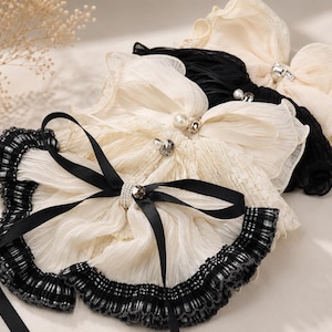 May include: A collection of layered hair accessories featuring pleated fabric bows in cream and black. The bows are adorned with central faux pearl and crystal embellishments, finished with a black ribbon tie and a ruffled black plaid trim edge.