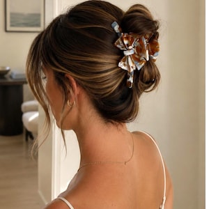Pu&ograve; includere: Un fermaglio per capelli con motivo tartarugato multicolore nei toni del marrone, bianco e blu. Il fermaglio ha un design a forma di fiocco con finitura lucida ed &egrave; fissato nei capelli castano scuro, raccolti in uno chignon morbido.