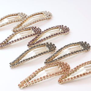 May include: Six hair clips adorned with rhinestones in shades of clear, purple, grey, and rose gold. The clips have a gold-coloured metal base and are arranged on a white surface. These accessories are perfect for adding sparkle to any hairstyle.