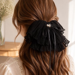 May include: A black hair accessory featuring layered, pleated fabric ruffles and a central decorative accent with a small white pearl and a clear crystal. The accessory includes thin black ribbon ties that hang down over the hair.