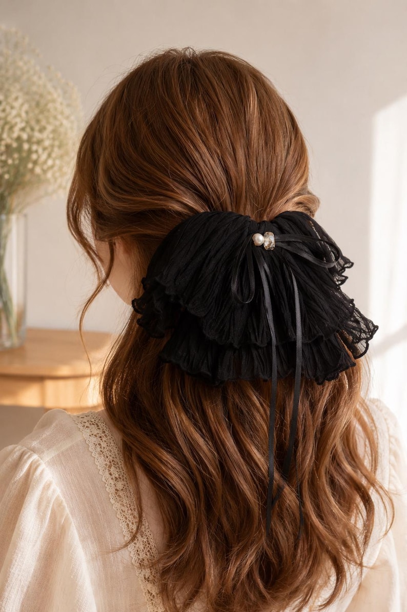 May include: A black hair accessory featuring layered, pleated fabric ruffles and a central decorative accent with a small white pearl and a clear crystal. The accessory includes thin black ribbon ties that hang down over the hair.