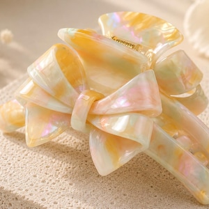 Pu&ograve; includere: Un fermaglio per capelli a molla con un fiocco decorativo sulla parte superiore. Il fermaglio &egrave; realizzato in materiale acrilico traslucido e marmorizzato con una delicata palette di colori giallo pallido, rosa chiaro e bianco, con una molla dorata.