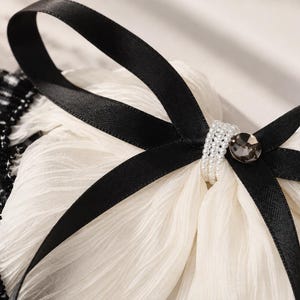 May include: A white pleated fabric accessory featuring a black satin ribbon tied into a bow. The center of the bow is embellished with a band of small white faux pearls and a single round dark gray faceted crystal. A black and white plaid trim.