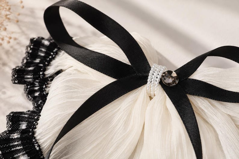 May include: A white pleated fabric accessory featuring a black satin ribbon tied into a bow. The center of the bow is embellished with a band of small white faux pearls and a single round dark gray faceted crystal. A black and white plaid trim.