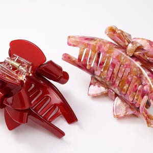 Pu&ograve; includere: Due pinze per capelli con design a fiocco. Una pinza &egrave; di colore rosso lucido, mentre l'altra presenta un motivo marmorizzato multicolore nelle tonalit&agrave; del rosa, bianco e giallo. Entrambe le pinze hanno un meccanismo a molla con denti.