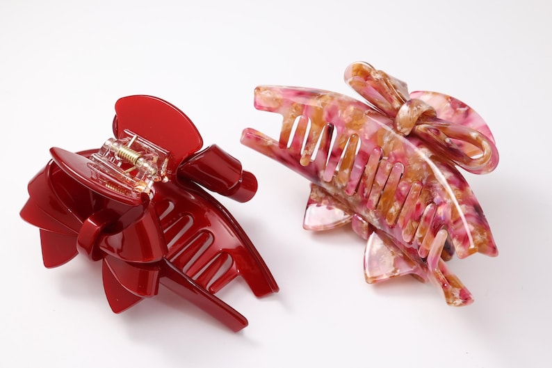Pu&ograve; includere: Due pinze per capelli con design a fiocco. Una pinza &egrave; di colore rosso lucido, mentre l'altra presenta un motivo marmorizzato multicolore nelle tonalit&agrave; del rosa, bianco e giallo. Entrambe le pinze hanno un meccanismo a molla con denti.