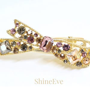 May include: Gold-toned hair clip in a bow shape, adorned with an array of sparkling crystals in shades of pink, gray, and clear. The clip features the text "ShineEve".